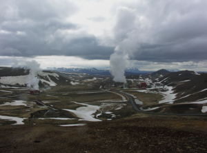 Steam rises from a the Krafla powerplant, a pair of small red buildings set in a vally. The landscape resembles a tundra, with patches of snow and a low range of mountains in the background