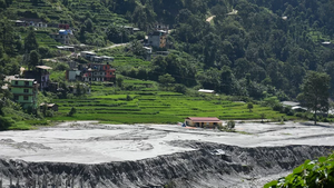 The foreground is filled with grey water as a result of flooding. In the middle of it sits a short building with a red roof and yellow walls. Increasing in elevation behind the flat ground affected by the flooding is a terraced field full of short, green vegetation and multi-storey buildings along the sides in a variety of colours. The right side towards the background is full of dark green trees.